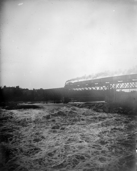 View upriver towards a train passing over a bridge. Smoke is trailing behind the locomotive, and a man is standing on top of the first railroad car.