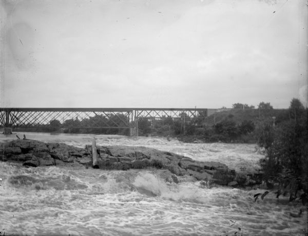 View upriver towards a railroad bridge over a river. A log is lying on a rocky protrusion in the river, and the far shoreline is on the right.