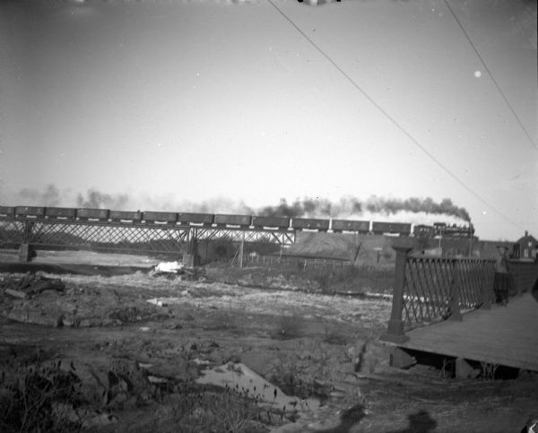 View towards a girl standing on a bridge on the right in the foreground. In the background a train is passing over a railroad bridge over a river. The bridge on the right is identified as the Harrison Street bridge in Black River Falls.