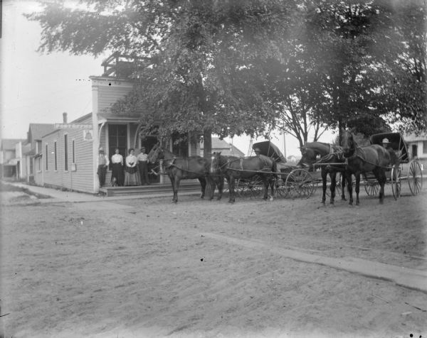 View towards two men and two women posing standing on the porch of a wooden store building on a city street, with three wagons and a horse on the right. Location identified as Warrens in Monroe County.