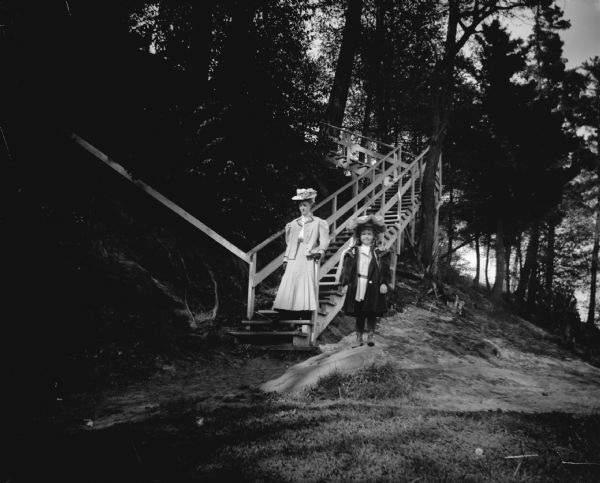 Outdoor portrait of a woman and a girl posing standing at the foot of steep wooden stairs on a tree-covered hill.