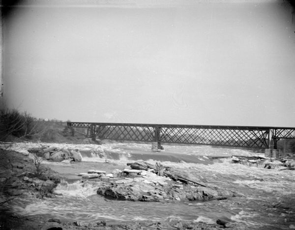 Elevated view upriver towards a railroad bridge crossing the river. Rocks are in the foreground in the middle of the river. Identified as the bridge across the Black River.