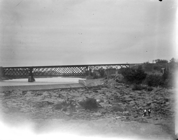 Elevated view upriver towards a bridge and dam. Two people are walking among the rocks on the lower right. Identified as the railroad bridge across the Black River.