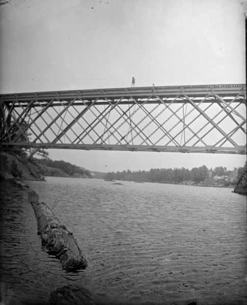 View across water towards two men posing standing on a railroad bridge across a river. Logs are floating in the water in the foreground.