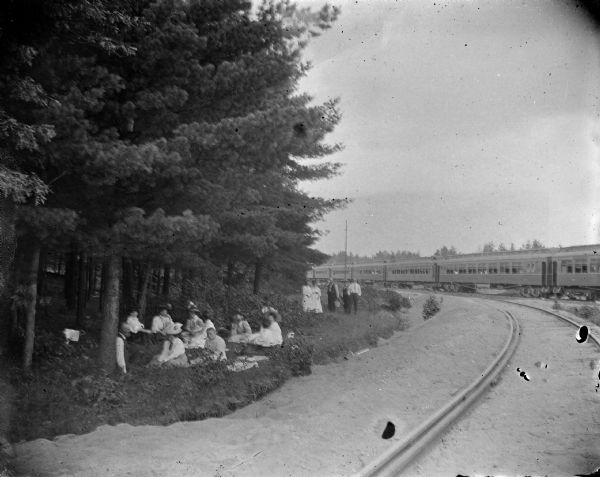 View down curving railroad tracks towards a group of men and women sitting and standing on the edge of a forest on the left. There is a passenger train on the tracks in the background on the right.