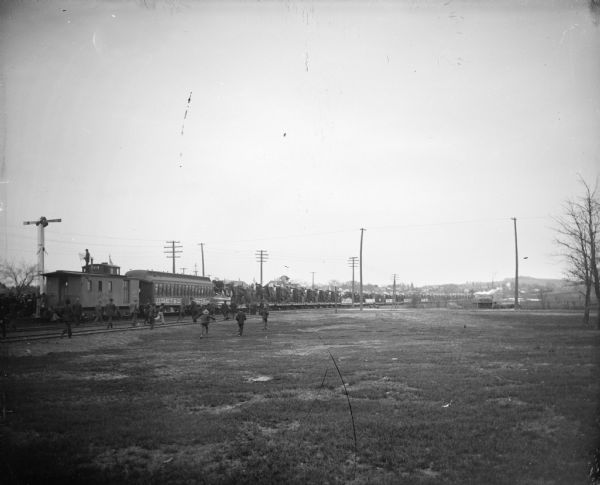View across open ground towards a group of men and boys moving towards a train. The train is identified as the train for J.T. Case.