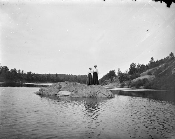 View across water towards two women posing standing on a rock in a river. Behind them are steep shorelines with trees and shrubs.