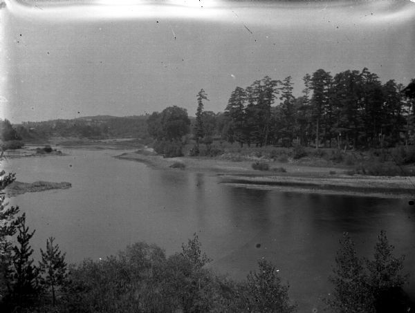 Elevated view down hill towards the tree-lined far shoreline along a river.