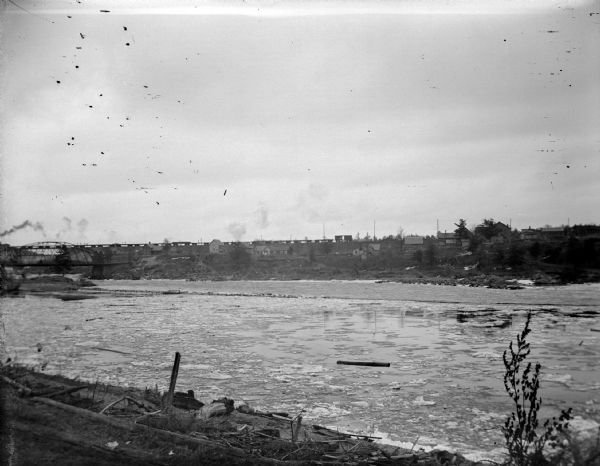 View from shoreline of an icy river towards a town on the opposite shoreline. There is a bridge crossing the river on the far left. Identified as the Black River showing the eastern shore.
