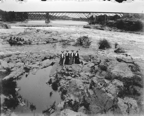Outdoor portrait from above of four women posing standing on rocks on a river. Another group of people are on the rocks behind them on the left. There is a railroad bridge across the river in the background.