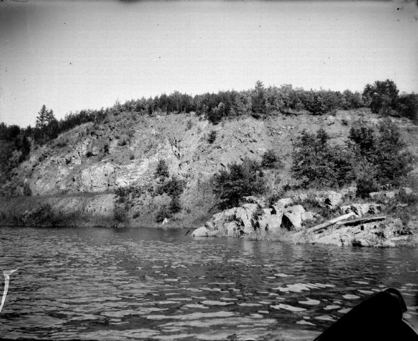 View across water towards the shoreline of a river. There are rocky outcroppings extending into the river, and the hill behind has exposed rock, with some trees along the base and along the top. Identified as the Black River.