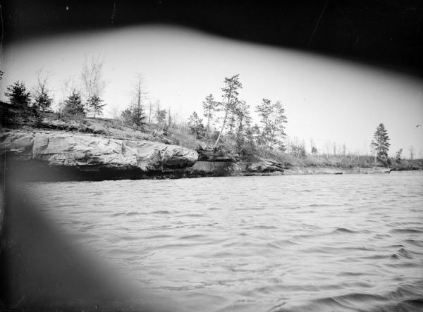 View across water towards the far shoreline of a river, with trees and plants growing on rocky outcroppings over the water.