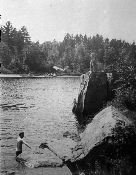 Copy photograph of an elevated view towards a nude man standing on a rock bluff along a river, and two other men in the water below and on the left. Another man is diving into the river. Identified as the swimming hole in the Black River just south of the cemetery at Spaulding Rock before the damming of the river.
