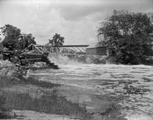 View from shoreline towards three men posing standing on wooden pilings next to a torrent of surging water of a river. There is a bridge over the river in the distance. Identified as the west channel of the Black River near a powerhouse in Black River Falls.