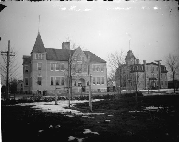 View across lawn of two large brick buildings. There are patches of snow on the ground. Identified as schools in Black River Falls, with the building on the left built in 1897, and the building on the right built in 1871.