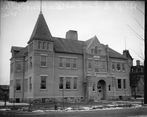 View across road towards a large brick building with patches of snow on the ground. Identified as the high school built in Black River Falls in 1897. Negative written on the emulsion side, "High School at Black River Falls, Wis."
