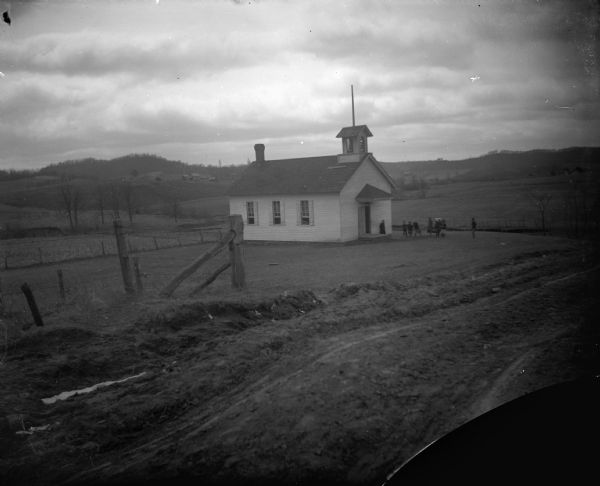 View down road towards of a small wooden school house with children in the yard in front of it. Identified as Shake Hollow school in the town of Franklin.