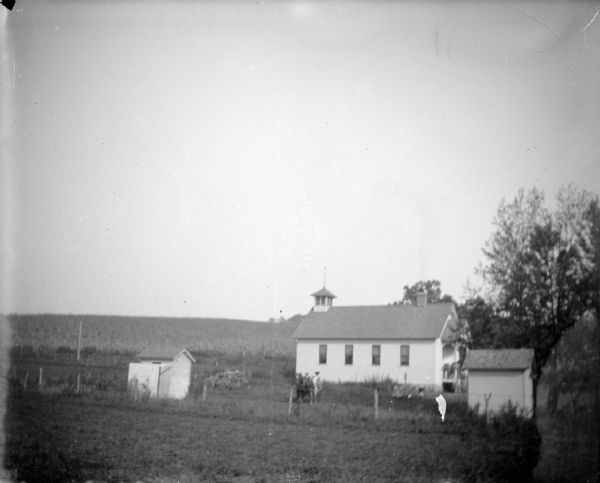 View across field towards a small wooden school house in the distance, with children on the porch, and a horse and wagon in the yard. Identified as the O'Brien school in the town of Irving.