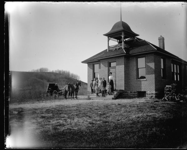 View across lawn towards a brick school house with children and a woman posing standing on the front steps. There is a man posing sitting in a wagon pulled by a team of two horses on the left. Identified as the school house in Kenyon.