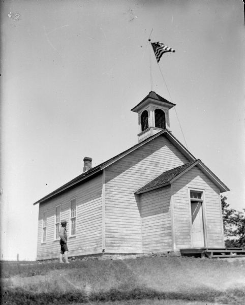View towards a boy posing standing in the yard of a wooden schoolhouse, which has a United States flag flying from the belltower. Identified as the school house in Disco.
