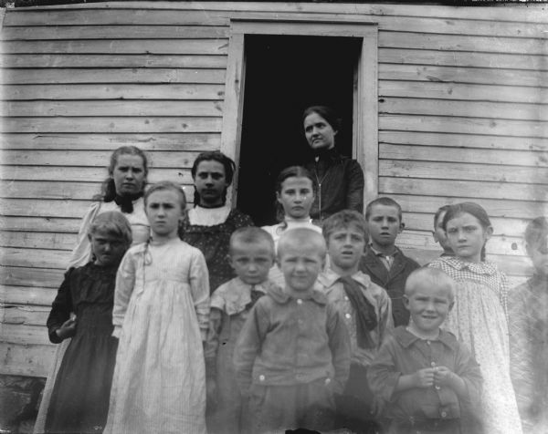Group portrait of a woman and children posing standing in front of the open doorway of a wooden building. Identified as a teacher and children at a rural school house.