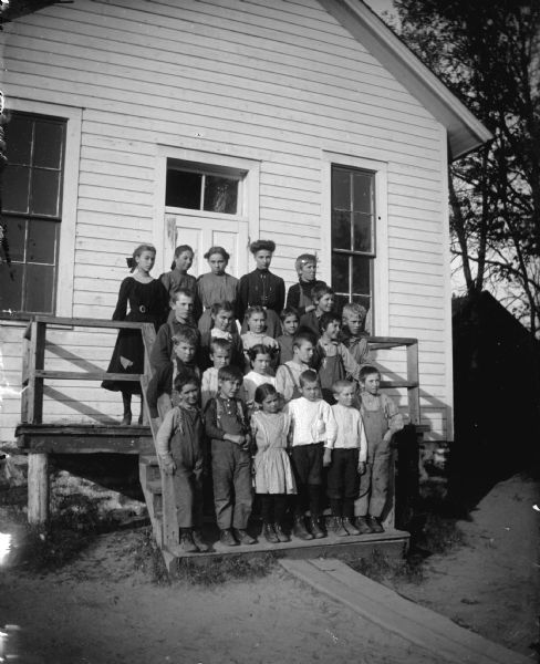 Group portrait of a woman and children posing standing on a wooden stairway of a wooden school house.