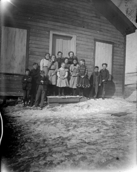 Group portrait of a woman and children posing standing on a wooden stairway of a wooden schoolhouse surrounded by snow-covered ground.