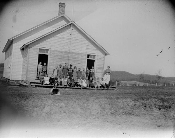 Outdoor group portrait of a woman and a large group of children posing standing and sitting on a wooden porch of a wooden school house with two open doors.