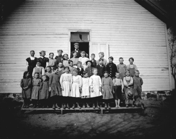 Outdoor group portrait of a man and a large group of children posing standing on the porch of a wooden schoolhouse.