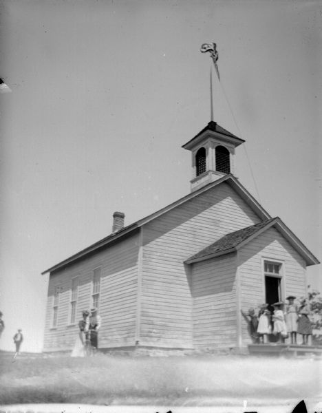 View across lawn towards a group of women, men, and children posing standing near and in front of a wooden schoolhouse.