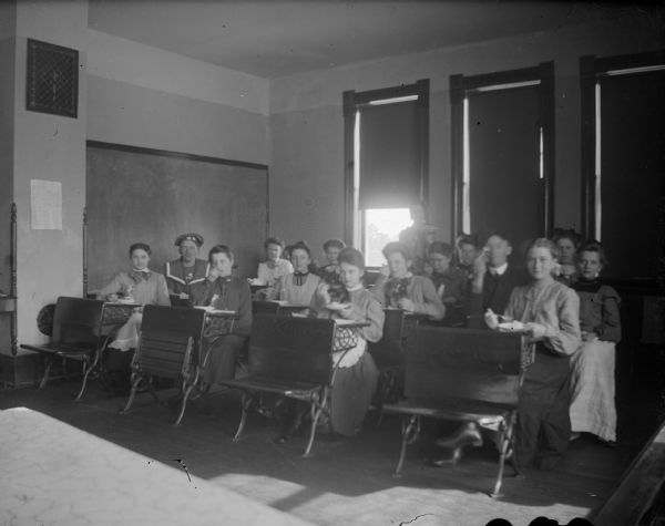 Indoor group portrait of men and women posing sitting in a classroom and eating. Identified as a classroom in the high school building built in 1897.