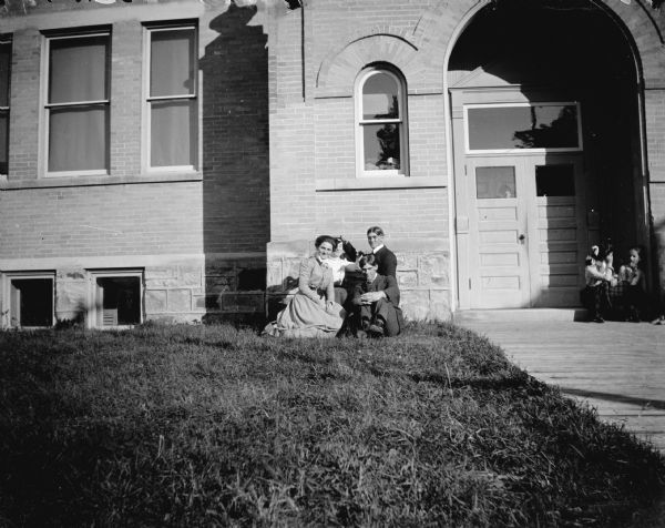 View across lawn and sidewalk towards of two men and two women posing sitting and kneeling in front of a large brick building. Another group is sitting near the front door. The building is identified as the Union High School built in 1871 in Black River Falls.