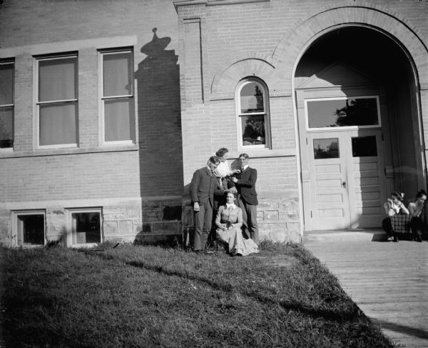 View across lawn towards two men posing standing and two women posing sitting and kneeling in front of a large brick building. Two women are sitting on the right in front of the arched entrance of the building. The building is identified as the Union High School built in 1871 in Black River Falls.