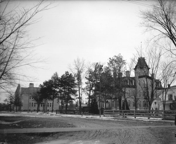 View down unpaved street towards two large brick buildings. There are patches of snow on the ground. Identified as the schools in Black River Falls.