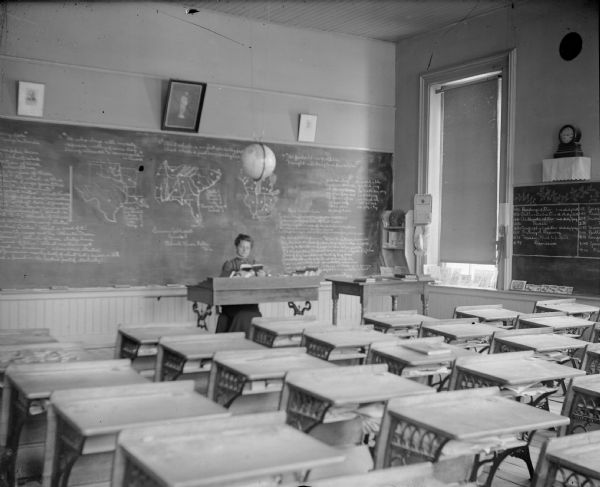 View from rear of classroom towards a woman posing sitting at a desk in front of a classroom. Identified as a room in the school building built in 1872 in Black River Falls.