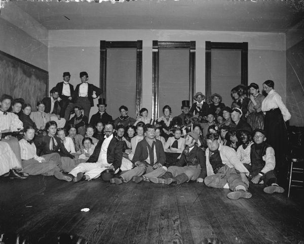 Interior group portrait of a large group of children and young adults posing standing and sitting in a room. Some of them are dressed in theatrical costumes and makeup. Identified as a classroom in the school building built in 1871.