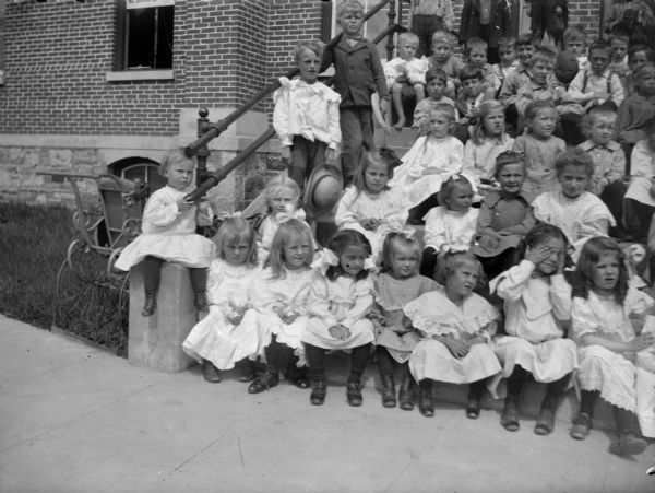 Outdoor group portrait of a large group of children posing sitting on a steps in front of a brick building.
