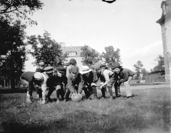Outdoor group portrait of nine boys crouched around a ball on a grass lawn in front of a large brick building.