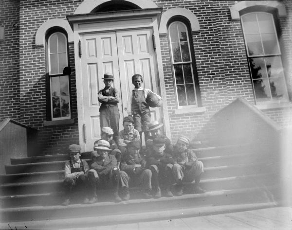 Group portrait of boys posing sitting and standing on a stairway in front of a brick building, which is identified as the school building built in 1872.