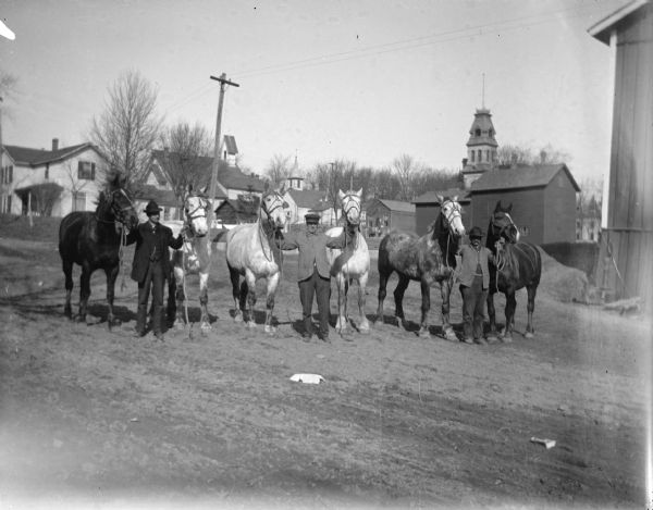 Outdoor group portrait of three men posing standing with six horses. Each man is standing between two horses and holding the reins. The location is identified as the intersection of Third and Fillmore Streets looking northwest.