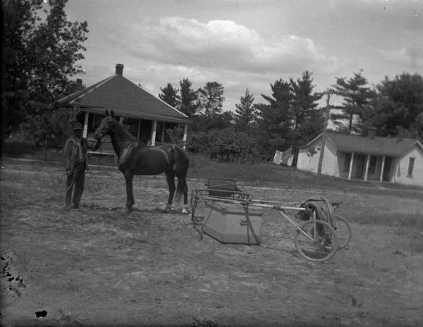 View towards a man displaying a single horse near a racing buggy resting on a trunk. There are buildings among trees in the background.