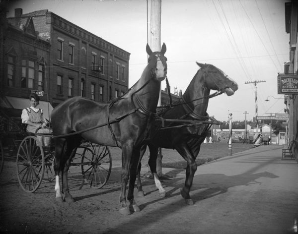 View towards two horses hitched to a wagon on a city street near a sidewalk. A woman is sitting in the wagon. The location is identified as looking east at the intersection of First and Main Streets. There is a barber's pole on the sidewalk in the background on the right.