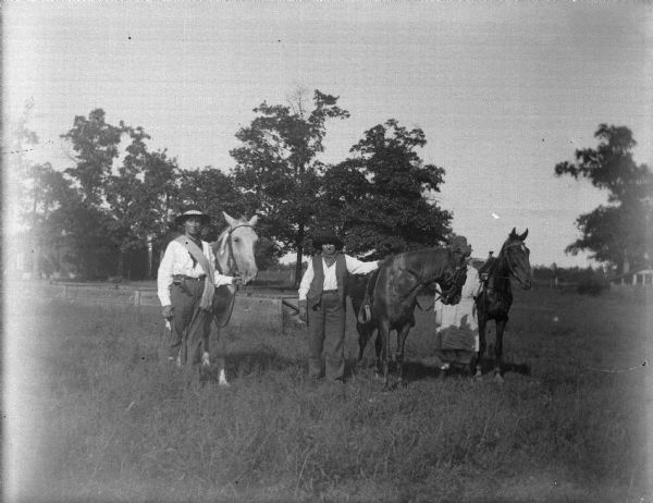 Outdoor group portrait of three women posing standing in a field. The two women on the left and center are wearing trousers and hats. The woman on the far right, whose face is obscured by one of the horses, is wearing a dress.