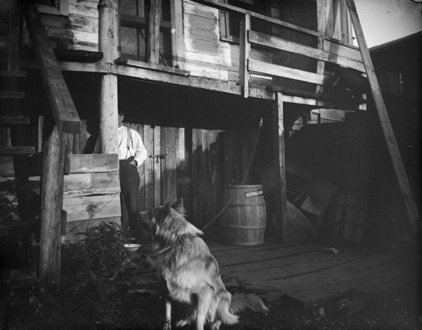 View towards a dog sitting at the end of a leash attached to the porch roof of a wooden building. Under the porch is a man, whose face is obscured by a column, standing near a closed door.