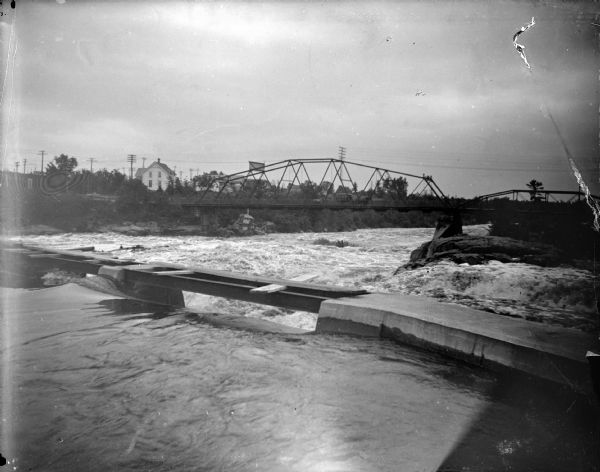 View downriver towards dam, with the water flowing under a bridge in the background. The bridge is identified as the wagon bridge, which was reportedly built in 1886 and removed in 1923 from over the Black River.