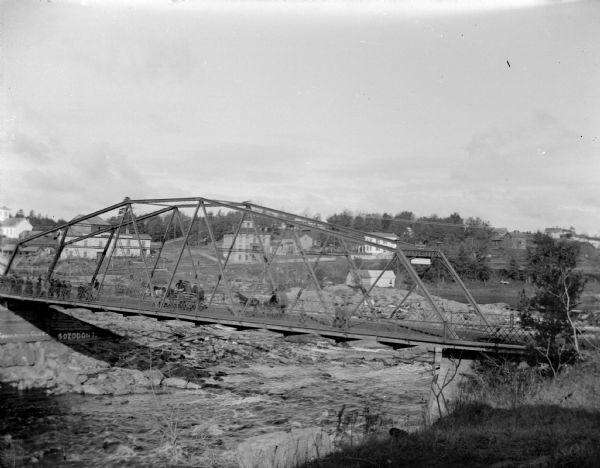 View from hill looking down towards individuals and wagons on a bridge over a river. The bridge is identified as the wagon bridge, which was reportedly built in 1886 and removed in 1923 from over the Black River.