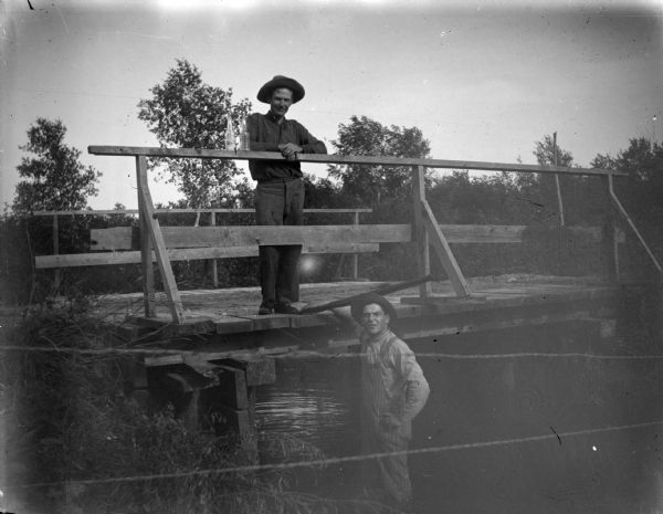 View through wire fence towards two men standing at a bridge. One of the men is standing on the bridge resting his elbows on the bridge railing. Two clear glass bottles are sitting on the railing next to him. The other man is standing on the riverbank just below the man. 