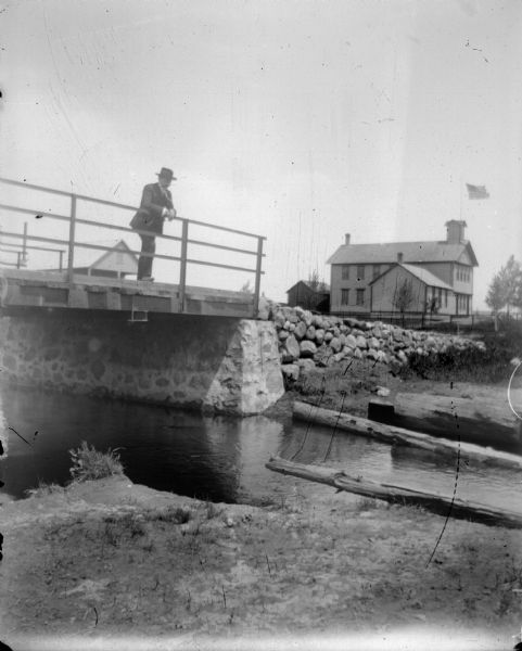 View from riverbank towards a man standing and leaning on the railing of a bridge. In the background is a large wooden building with a flag on the roof.