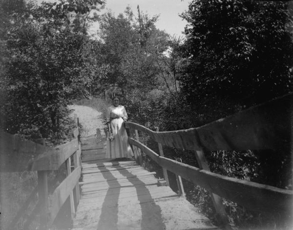 View down wooden foot bridge towards a woman standing with her arm resting on the railing. She is holding a bunch of flowers in her right hand. In the background is a pathway leading to the bridge.