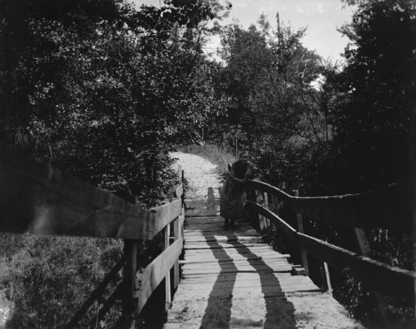 View down wooden foot bridge towards a woman standing and leaning on a the railing. There is a pathway in the background leading to the bridge.